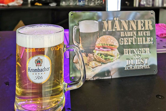 Gefülltes Bierglas mit einem Aluschild im Hintergrund. Auf dem Schild steht "Männer haben auch Gefühle! Hunger und Durst zum Beispiel."